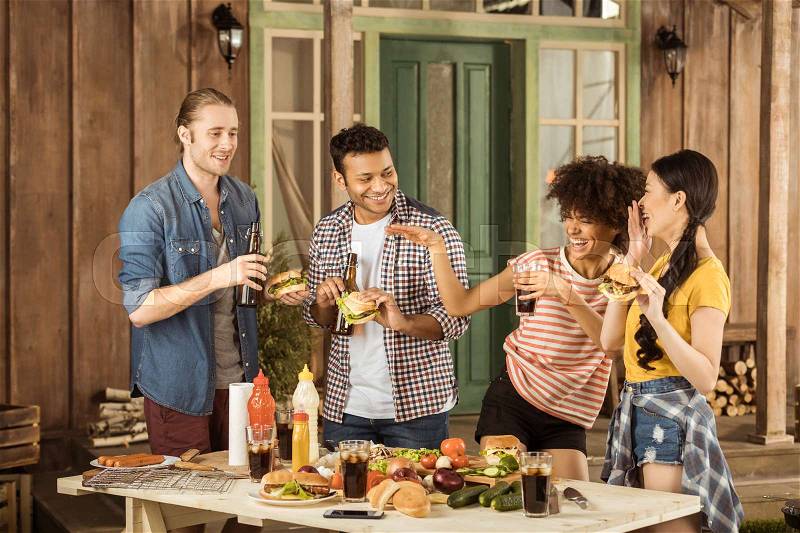 Young multiethnic friends fooling around and eating burgers at picnic on patio, stock photo