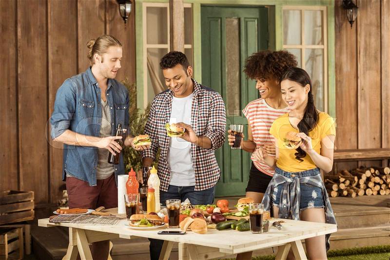 Young multiethnic friends eating burgers and drinking cola at picnic on patio, stock photo