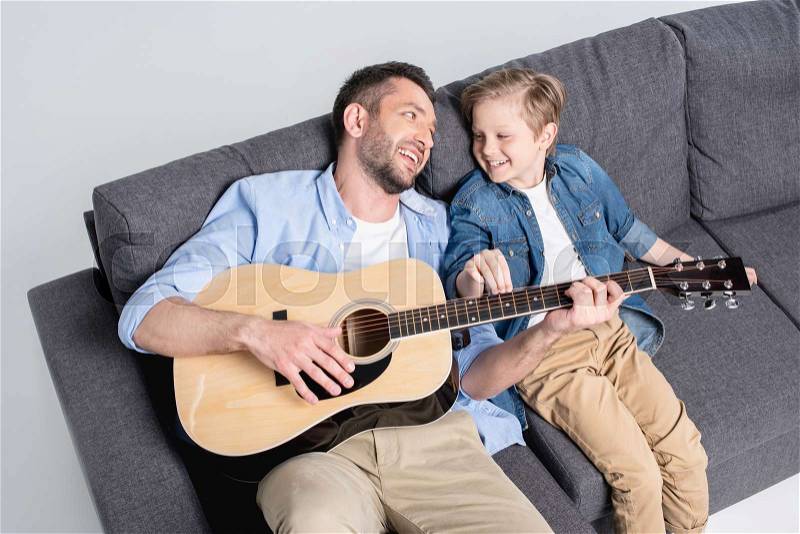 Father playing on guitar and looking on his son while sitting on sofa, stock photo