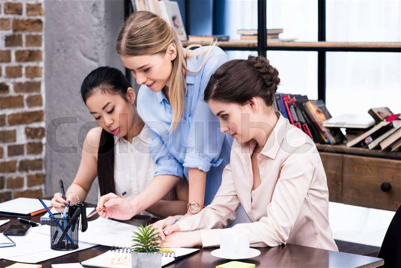 Multiethnic group of young businesswomen working together at table with papers, stock photo
