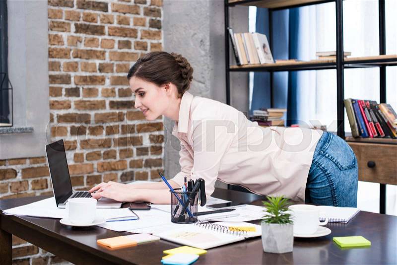 Smiling young woman using laptop at desk with papers and office supplies , stock photo