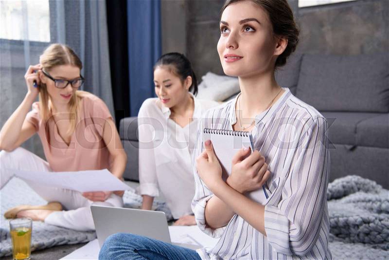 Young woman dreaming and holding notebook while her colleagues working at home, stock photo