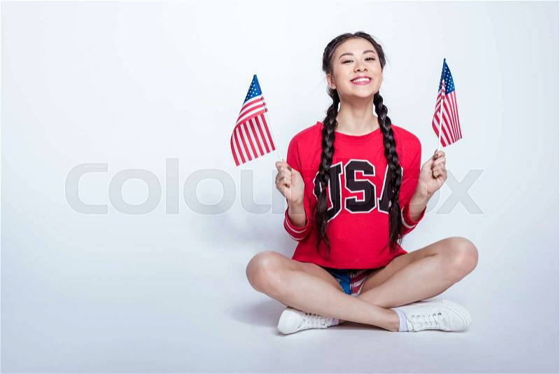 Happy asian girl in red sweatshirt with USA word sitting and holding little american flags isolated on grey, America\'s Independence Day concept, stock photo