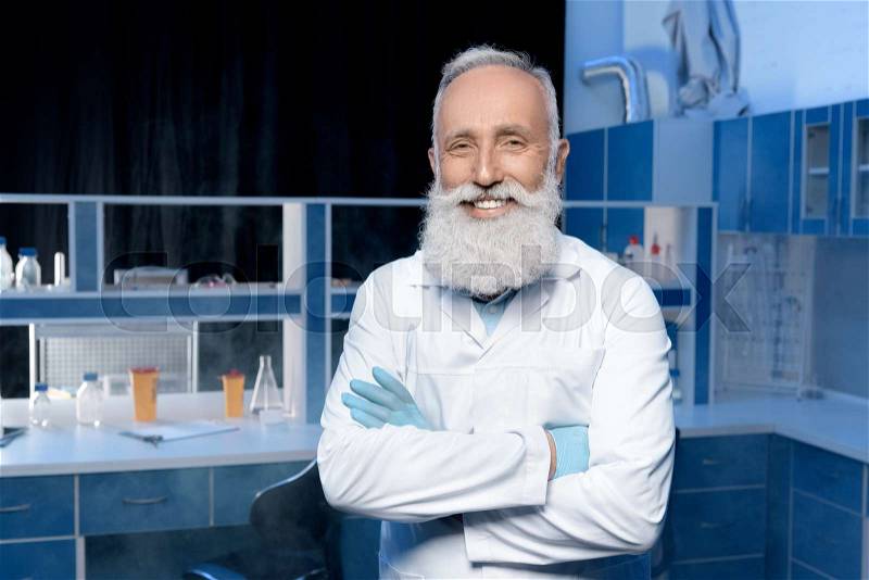 Smiling grey haired scientist in lab coat looking at camera with arms crossed, stock photo