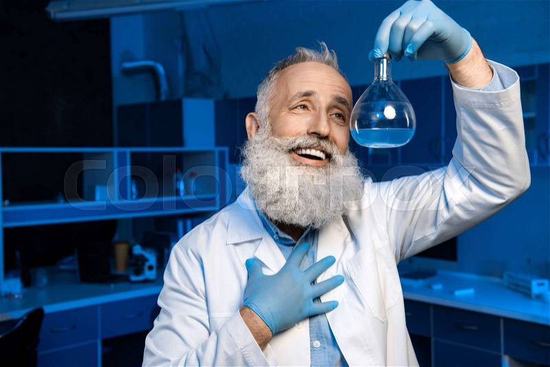 Happy grey haired scientist in lab coat holding flask with reagent, stock photo