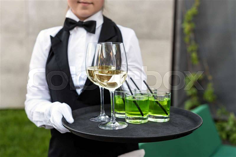Midsection of professional waiter in uniform serving wine during buffet catering party, festive event or wedding. Full glasses of champagne on tray. Outdoor party catering service, waiter on duty, stock photo
