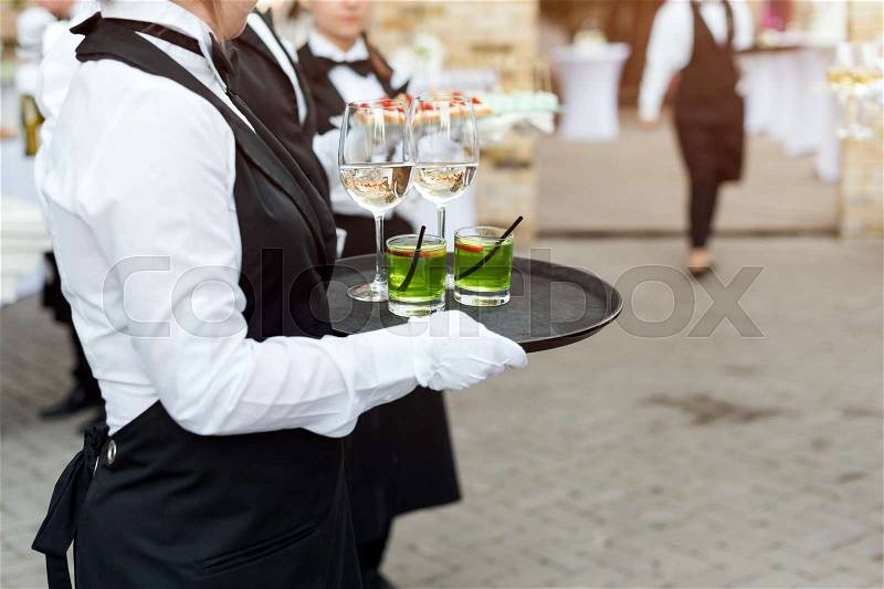 Midsection of professional waiters in uniform serving wine, cocktails and snacks during buffet catering party, festive event or wedding. Full glasses of wine on tray. Outdoor party catering service, stock photo