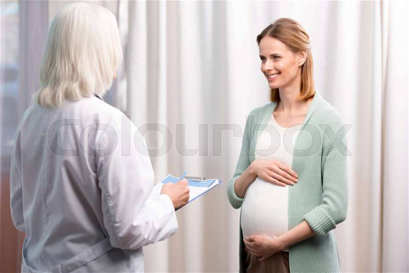 Doctor writing down young pregnant woman complaints during medical consultation, stock photo