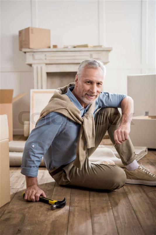 Smiling senior man sitting on floor with hammer in hand at new home, stock photo