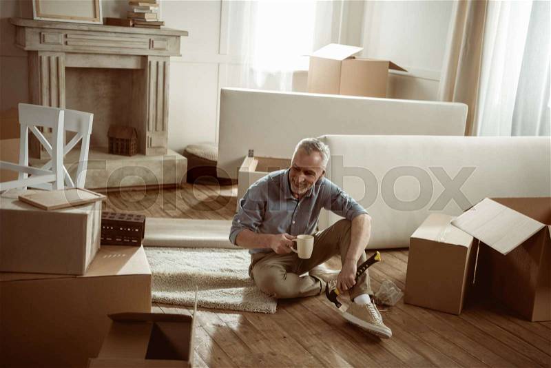 Happy senior man sitting on floor with cup of tea and taking break from packing cardboard boxes, moving out concept , stock photo