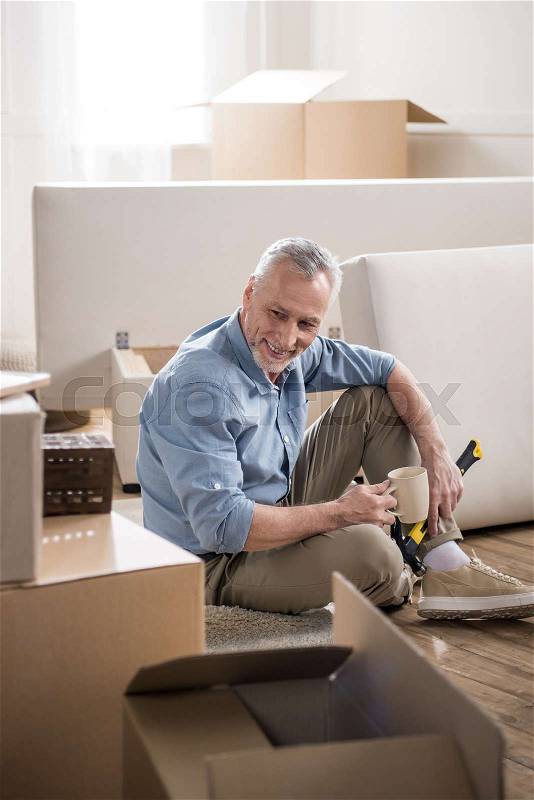 Casual senior man sitting on the floor with hammer and drinking coffee at home, stock photo