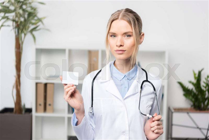 Confident young doctor holding blank business card and looking at camera, stock photo