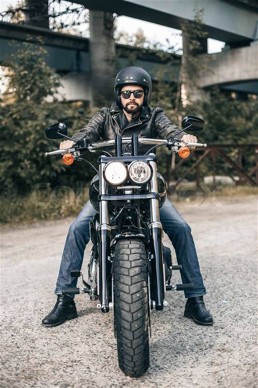 Handsome rider guy with beard in black biker jacket, sunglasses on classic style cafe racer motorcycle at sunset. Brutal fun urban lifestyle, stock photo