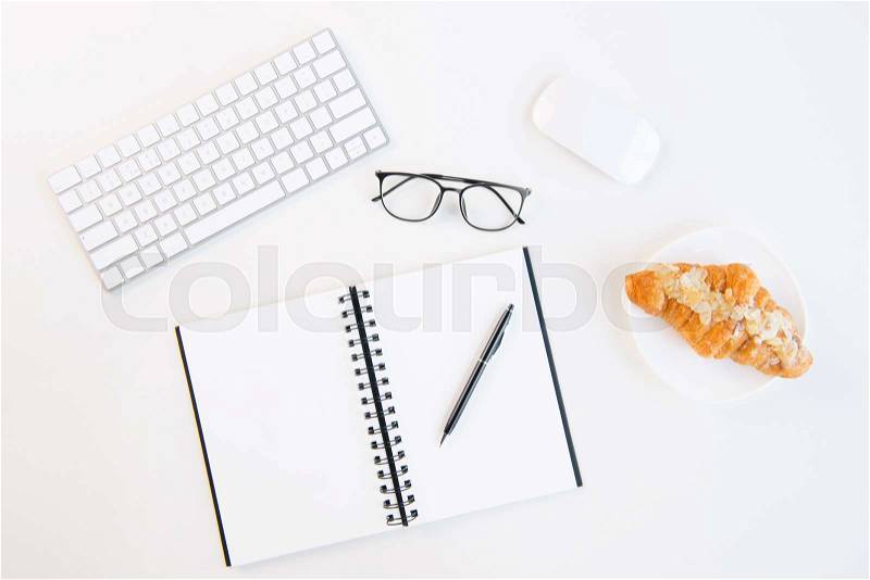 Top view of tasty croissant on plate, blank notebook with pen, keyboard and computer mouse at workplace, stock photo