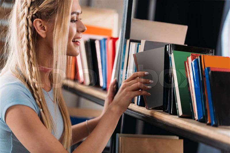 Smiling young woman choosing book on bookshelf in library , stock photo