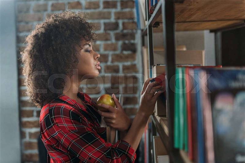 Side view of beautiful african american student holding books with apple and looking at bookshelves , stock photo