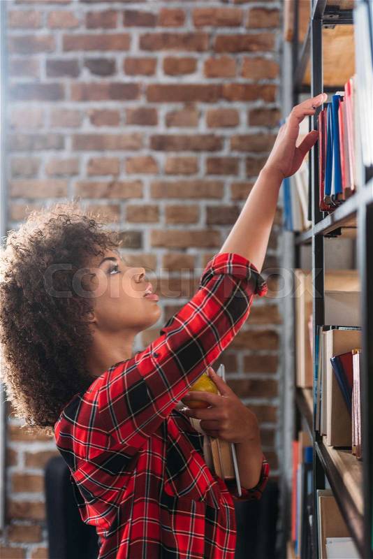 Side view of beautiful african american student holding books with apple and looking at bookshelves , stock photo