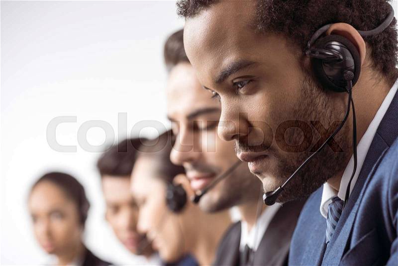 Close-up view of focused call center operator in headset working with colleagues behind, stock photo