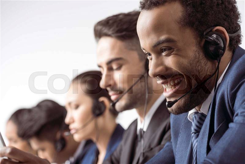 Close-up view of smiling call center operator in headset working with colleagues behind , stock photo