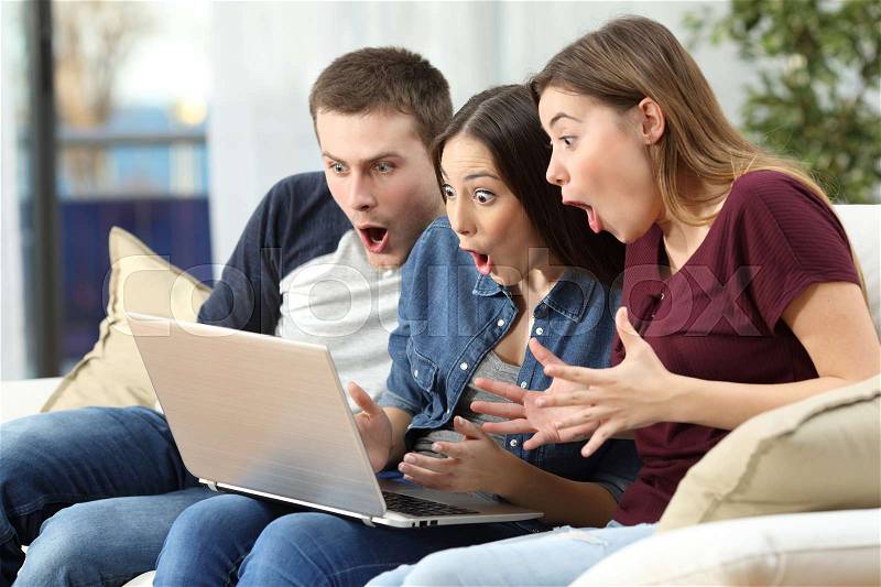 Three amazed friends watching media content on line in a computer sitting on a couch in the living room at home, stock photo