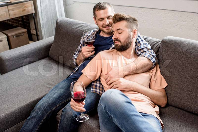 Casual homosexual couple embracing and drinking wine while on sofa at home, stock photo