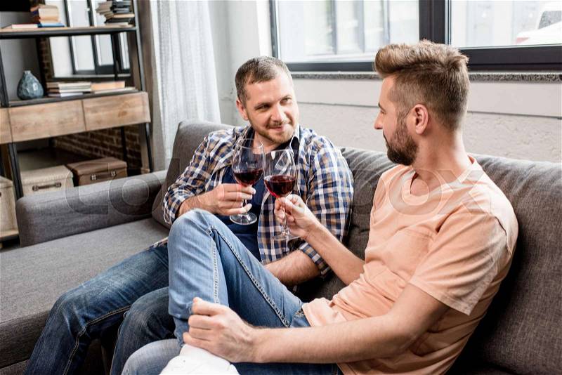 Smiling homosexual couple clinking with glasses of wine while sitting on sofa at home, stock photo