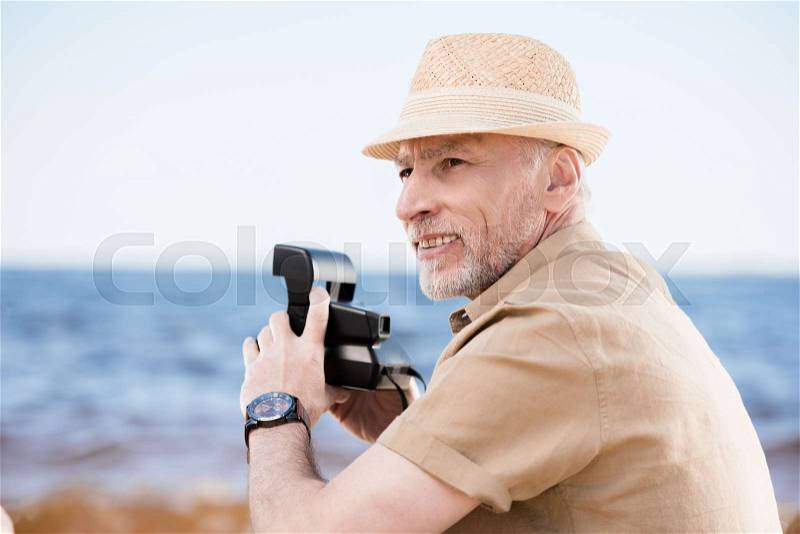 Smiling senior man in hat holding instant camera and looking away outdoors, stock photo