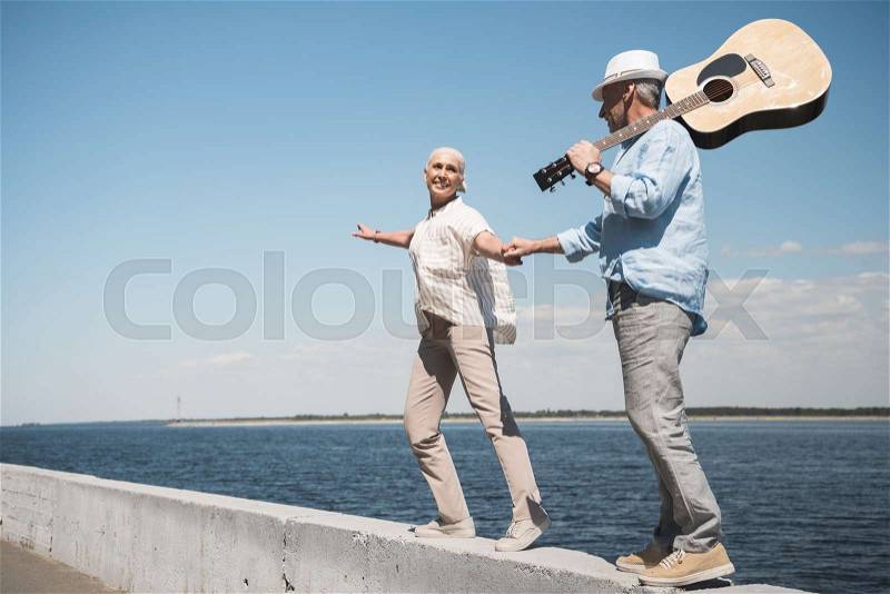 Happy senior couple with guitar holding hands and walking at quay, stock photo