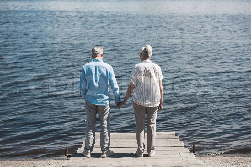 Back view of senior couple standing and holding hands on riverside at daytime, stock photo