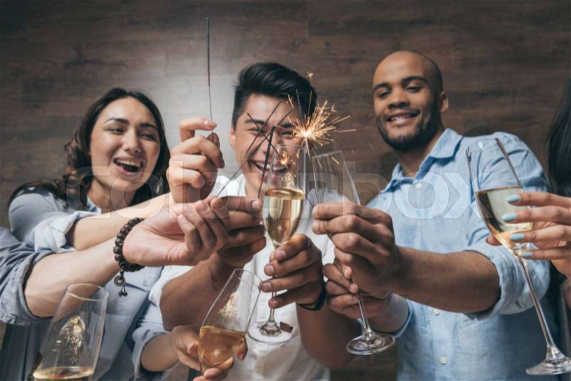 Cheerful young people holding glasses of champagne and burning sparklers indoors , stock photo
