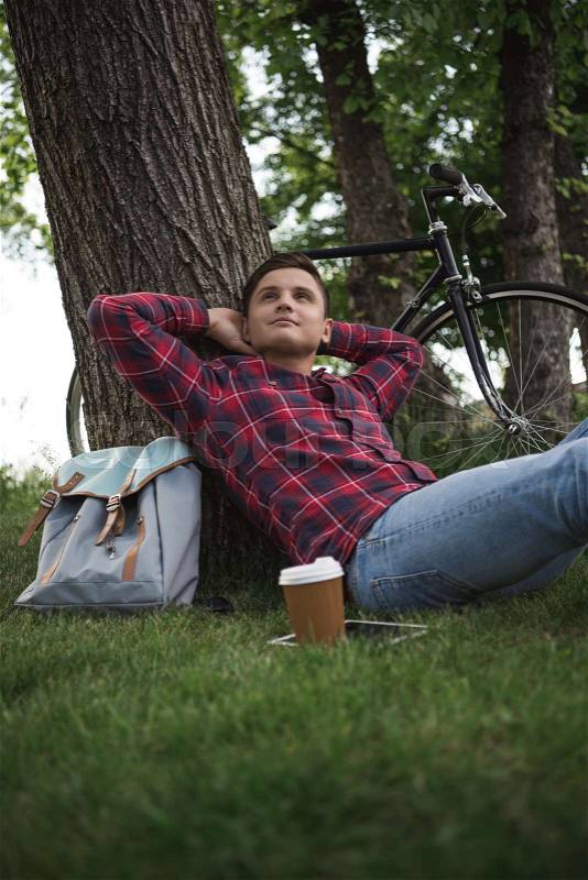 Young man enjoying summer day with coffee cup, digital tablet and bicycle behind of tree, stock photo