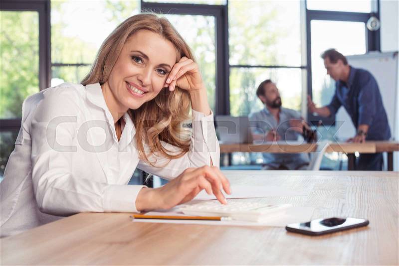 Middle aged businesswoman using calculator and smiling at camera, stock photo