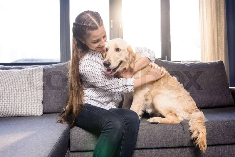 Teenager girl hugging golden retriever dog on sofa at home, stock photo