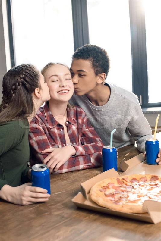 Side view of friends kissing smiling teenage girl\'s cheeks, eating pizza at home concept , stock photo