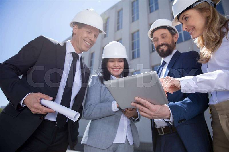 Smiling professional architects in hard hats holding blueprint and using digital tablet, stock photo