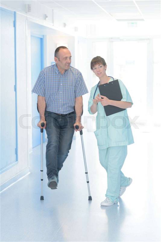 Nurse helping man with crutches in hospital corridor, stock photo