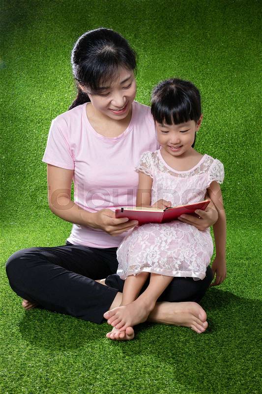 Asian Chinese mother and daughter sitting on the grass and reading the book at outdoor park, stock photo