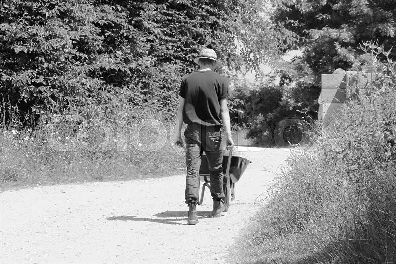 The male employee with straw hat is walking with the wheelbarrow over the terrain of Great Dixter House & Gardens in England in the beautiful summer in black and white, stock photo