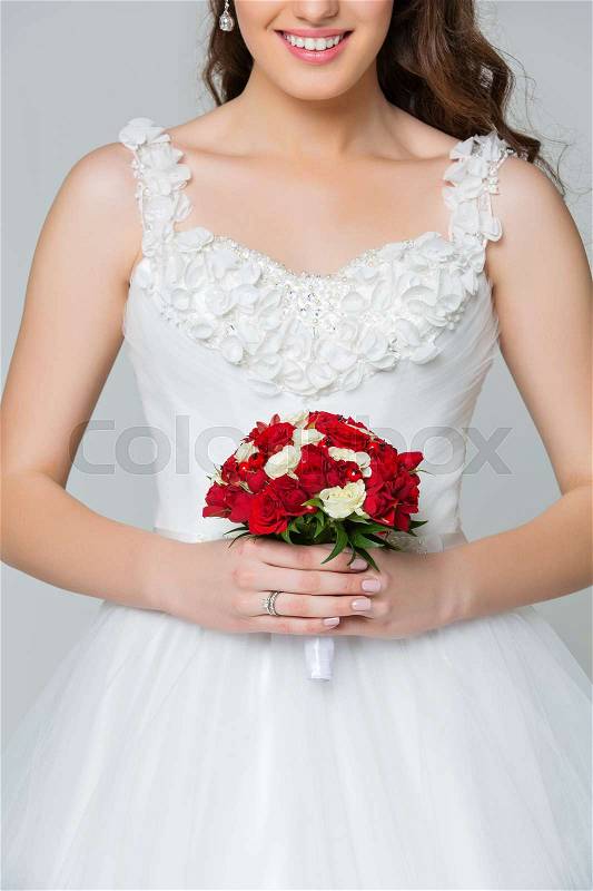 Beautiful happy young bride with red bouquet in long white wedding gown. Studio shot on grey background. Copy space, stock photo