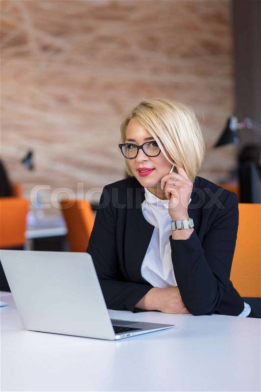 Good business talk. Cheerful young beautiful woman in glasses talking on mobile phone and using laptop with smile while sitting at her working place, stock photo