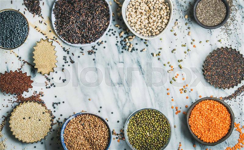 Various raw uncooked grains, beans, cereals in bowls and cups for healthy cooking over marble background, top view, copy space. Clean eating, dieting, healthy, detox, vegan, vegetarian food concept, stock photo