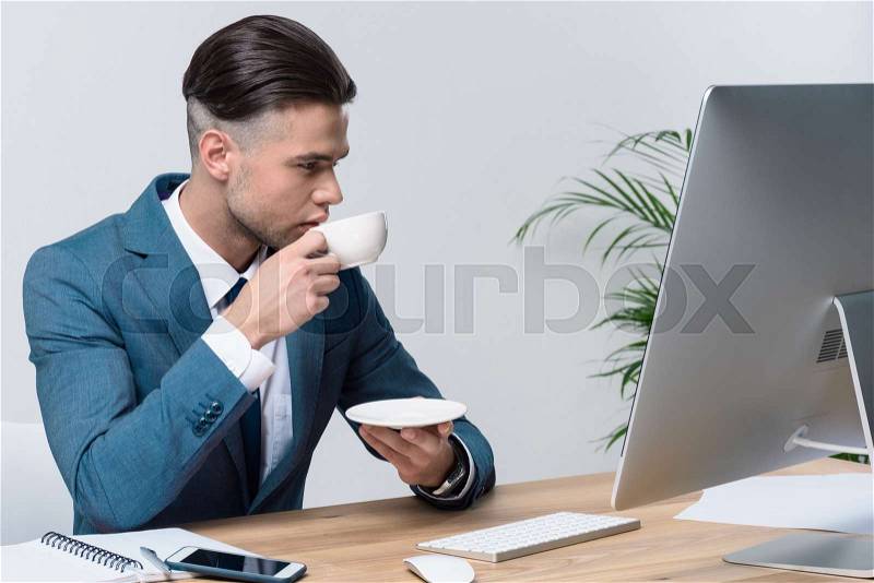 Handsome young businessman drinking coffee while using desktop computer in office, stock photo