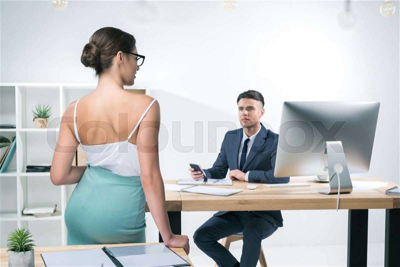 Handsome young businessman working at desk and looking at seductive businesswoman standing in office , stock photo