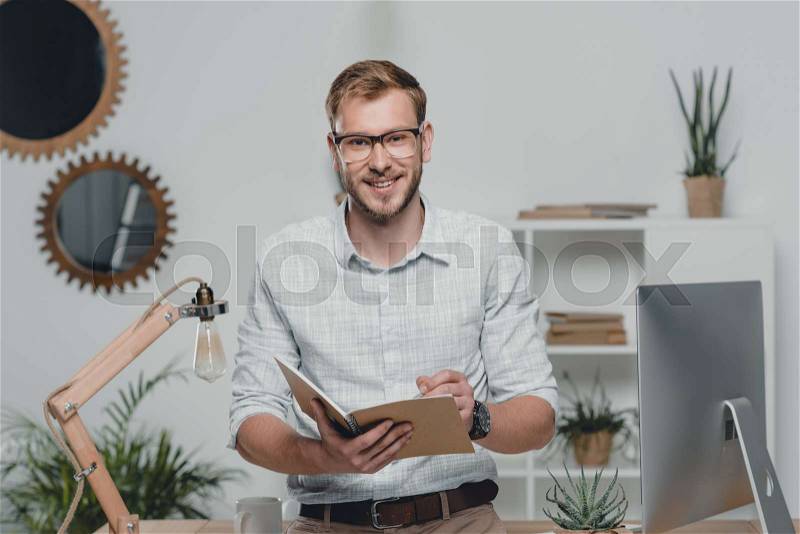Happy caucasian businessman writing in diary and smiling at camera in modern office, stock photo