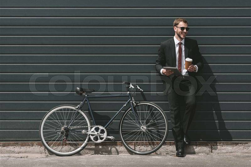 Stylish businessman with bicycle using digital tablet and drinking coffee while standing in front of black wall, stock photo