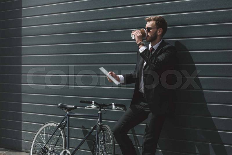 Stylish businessman with bicycle using digital tablet and drinking coffee while standing in front of black wall, stock photo