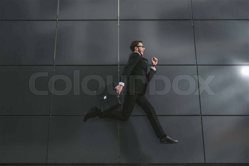 Excited stylish businessman in suit and sunglasses jumping in front of black wall, stock photo