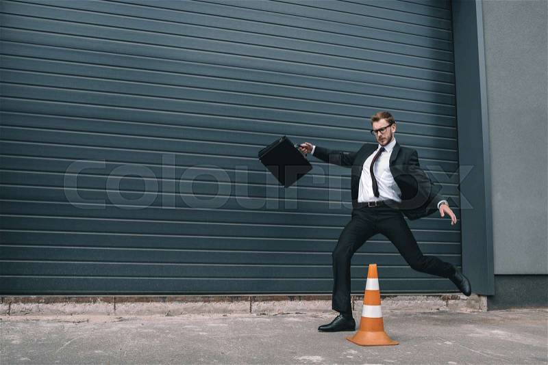 Stylish businessman in eyeglasses holding briefcase and running near traffic cone , stock photo
