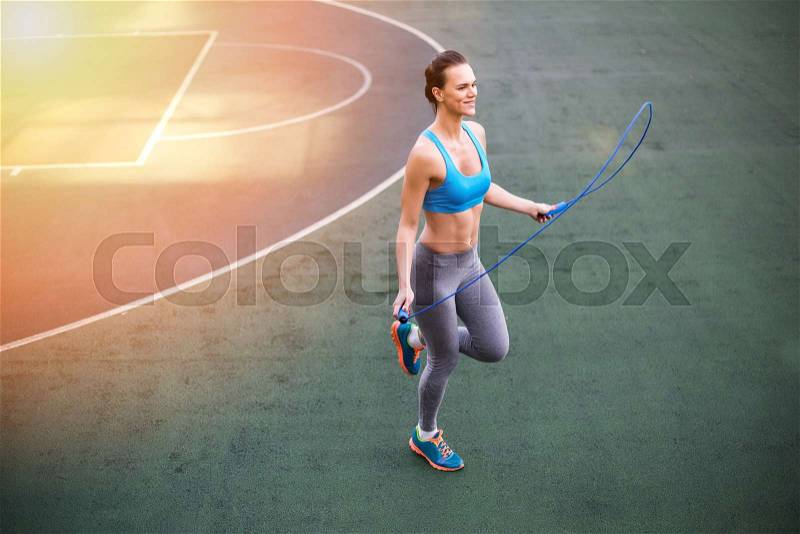 Smiling young woman in sportswear exercising with skipping rope on stadium, stock photo