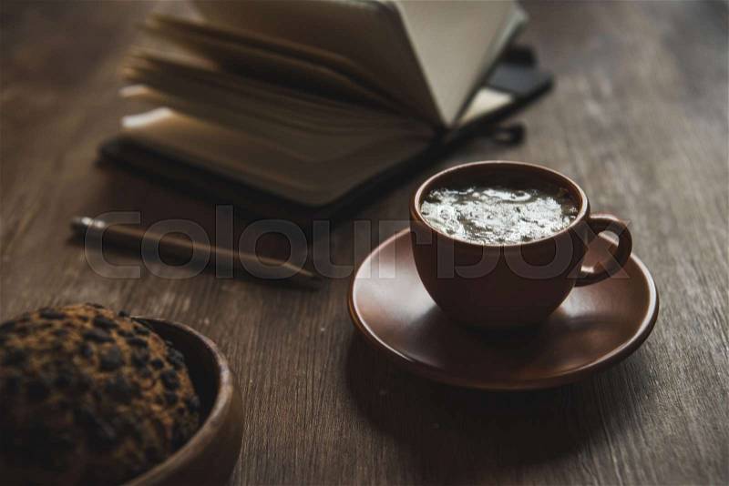 Close-up view of open notebook with pencil, cup of coffee and chocolate chip cookie on wooden table, stock photo